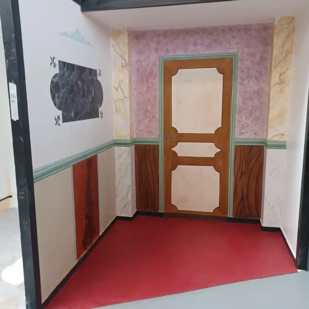 Cette photo d’un petit salon présente de larges panneaux imitant la veine du marbre ainsi que des moulures et portes en faux bois viennent structurer les murs et soulignent les différents motifs. Elles donnent l'impression d'une boiserie classique, ajoutant une touche chaleureuse et intemporelle. Des pochoirs de fleurs de lys  , ornent certains éléments architecturaux, tels que les encadrements, fronton ou corniches. Ces motifs apportent une note de légèreté et de romantisme.